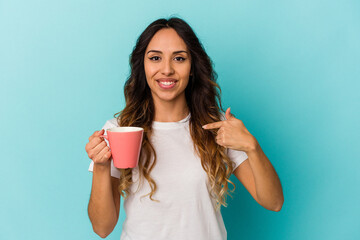 Young mexican woman holding a mug isolated on blue background person pointing by hand to a shirt copy space, proud and confident