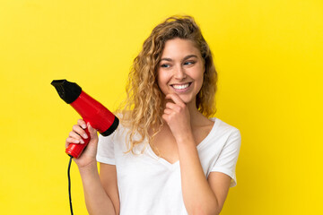 Young blonde woman holding a hairdryer isolated on yellow background looking to the side and smiling