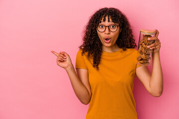 Young mixed race woman holding a cookies jar isolated on pink background pointing to the side