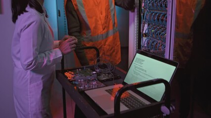 Slowmo tilt up shot of female Asian IT specialist in lab coat typing on laptop and checking computing equipment with male African-American technician in data center - Powered by Adobe