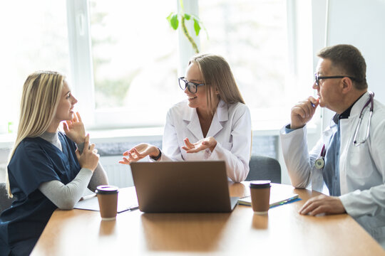 Multiracial Medical Team Having A Meeting With Doctors In White Lab Coats And Surgical Scrubs Seated At A Table Discussing A Patients Records