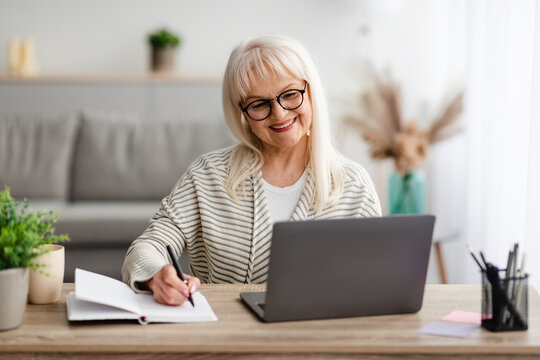 Mature woman in glasses writing and using laptop at home