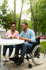 Happy disabled black man and his female coworker using laptop together at table in outdoor cafe, copy space