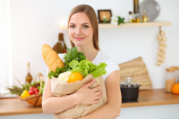 Obraz na płótnie Canvas An attractive young woman holding the paper bag full of vegetables while standing and smiling in sunny kitchen. Cooking concept