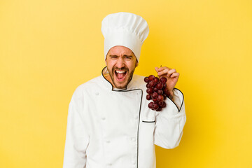 Young caucasian chef man holding grapes isolated on yellow background screaming very angry and aggressive.