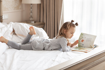 Child girl relaxing and reading interesting book stories in comfortable bed in the morning near...