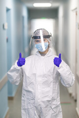 Fototapeta na wymiar Thumbs up in blue medical gloves. A girl doctor in a protective suit during an epidemic covid in the white corridor of a modern hospital.