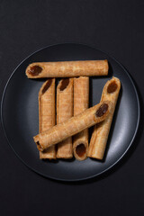 Photo of a dish with homemade waffle rolls with boiled condensed milk and nuts on a black plate, close-up. 