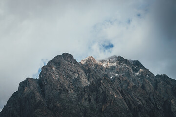 Atmospheric mountain landscape with low cloud on mountain top in sunlight. Dark rocks with snow and blue skylight in gray cloudy sky. Beautiful mountain scenery with low cloud on rocky top in sunshine