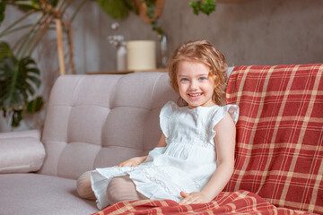 Happy cute little girl sitting at home on the couch smiling, looking at the camera