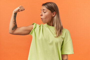 Surprised beautiful young woman in yellow tshirt showing biceps muscles and looking on it isolated over orange background - obrazy, fototapety, plakaty