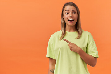 Amazed attractive young woman in yellow tshirt standing and pointing away to the side at empty space by finger isolated over orange background