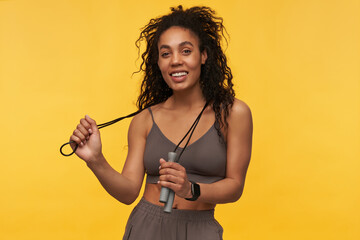 Portrait of cheerful beautiful african american young sportswoman standing and holding skipping rope on her neck isolated over yellow background