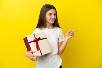 Little girl holding a gift over isolated yellow background pointing finger to the side