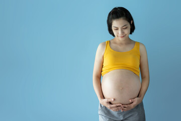 Portrait of Smiling Asian Pregnant Female standing touching her belly on blue isolated background