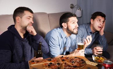 adult male friends in suspense watching tv together at home, drinking beer with pizza