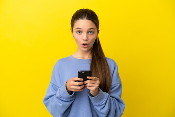 Little girl over isolated yellow background looking at the camera while using the mobile with surprised expression