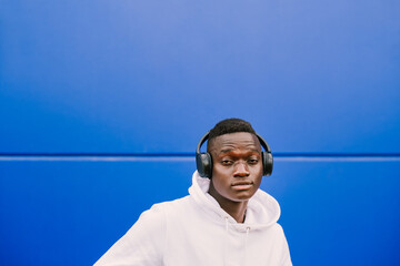 Young African American black man listening music with wireless headphones while wearing a white sweatshirt, looking to camera. Blue wall background