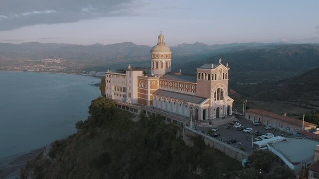 Aerial parallax shot of Sanctuary of Tindari baroque facade from high angle view at sunset, iconic Sicily travel destinations in Italy. Beautiful christian baroque cathedral by the coastline