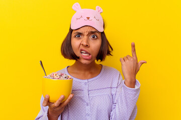 Young mixed race woman eating cereals wearing a pijama isolated on yellow background showing a...