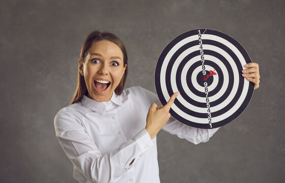 Wow, right in bullseye. Studio portrait of excited smiling lady looking at camera pointing finger at shooting target with red dart in center. Business goal, target market, objective, success concept