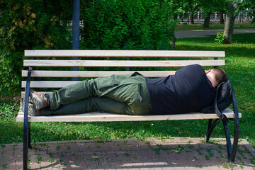 A tired tourist sleeps on a bench in the park, putting a backpack under his head.