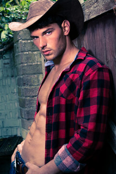 Handsome cowboy looking at camera wearing hat with open chequered shirt revealing defined pecs and sixpack abs