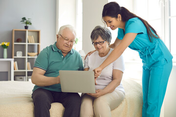 Smiling home care nurse teaching old patients to use modern computer. Happy senior couple looking at laptop screen and learning to make video calls, browse websites and visit online medical platforms