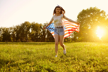 Independence Day USA. Girl with American flag runs on the grass in a summer park at sunset.