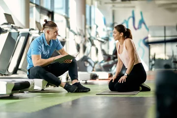 Schilderijen op glas Happy athletic woman talking to her personal trainer during break in a gym. © Drazen