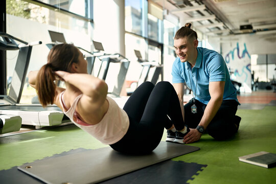 Happy male coach assisting sportswoman in exercising sit-ups in a gym.