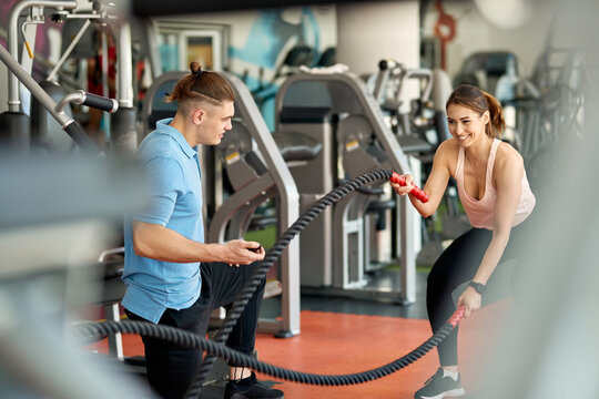Happy sportswoman exercising with battle ropes while working out with personal trainer in a gym.