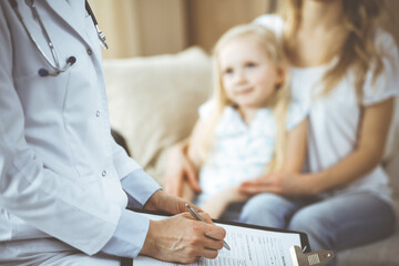 Fototapeta premium Doctor and patient. Pediatrician using clipboard while examining little girl with her mother at home. Happy cute caucasian child at medical exam. Medicine concept