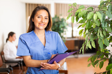 Positive Latino doctor woman standing in office with clipboard, writing medical history sheet
