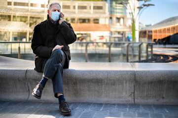 Senior man on the phone outdoor sitting on a bench and wearing a mask, coronavirus concept