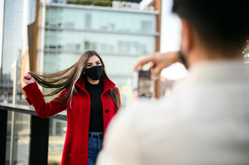 Man taking pictures of his girlfriend outdoor wearing protective mask against covid coronavirus pandemic