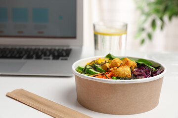 Delicious salad with chicken and vegetables in paper bowl near laptop on white table. Space for text