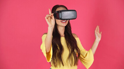 young adult woman in vr headset gesturing with pointing finger isolated on pink