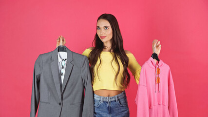 dreamy young adult woman holding hangers with blazer and hoodie isolated on pink