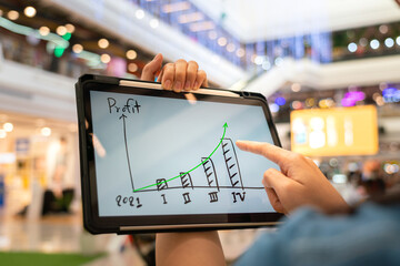 A manager using tablet to see business growth predictation graph of 2021 on screen with blur background of department store interior. Business successful concept. Selective focus at human hand part.