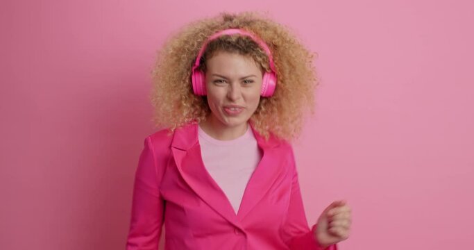 Joyful upbeat young woman keeps hands on her curly hair dressed in jacket listens audio track poses against pink background. Overjoyed European female feels very happy enjoys music in headphones