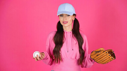 young adult woman in cap and hoodie holding baseball equipment isolated on pink