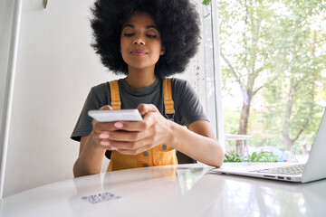 Closeup shot of young black gen z African American girl with afro hair holding cell smart phone scanning qr code to read menu or make payment online in cafe sitting at table indoors.