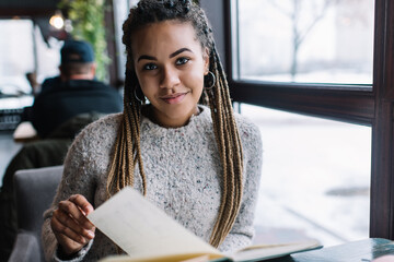 Portrait of charming hipster girl with knowledge notebook looking at camera during studying time,...