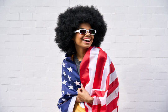 Happy smiling African American teenager wearing sunglasses wrapped in usa flag celebrating july 4th forth independence day of united states. Patriotic portrait on white bricks wall background.