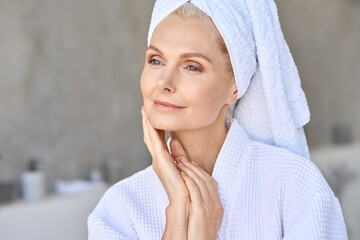 Happy beautiful middle aged woman wearing bathrobe and white towel with perfect complexion touching face looking away in bathroom. Advertising of skin care spa wellness concept. Closeup portrait. - obrazy, fototapety, plakaty