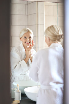 Vertical shot of attractive senior adult 50s blonde woman in bathroom wearing bathrobe touching face after washing and beauty morning routine looking at reflection in mirror smiling. Skincare concept.
