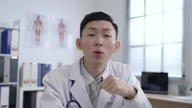 shoulder view asian physician facing camera is expressing with hand gestures and inviting others to speak while hosting a remote conference at clinic.