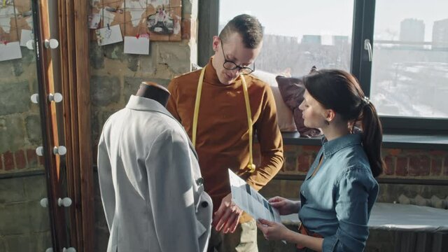 Medium shot of male fashion designer in glasses looking at garment hanging on mannequin in studio and talking to female seamstress showing him fabric samples