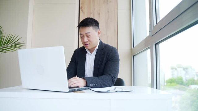 young asian business man sitting in the office talking online via webcam, looking at laptop. Male in a formal suit, an employee or manager waving his hand, happy talking on a video call indoors